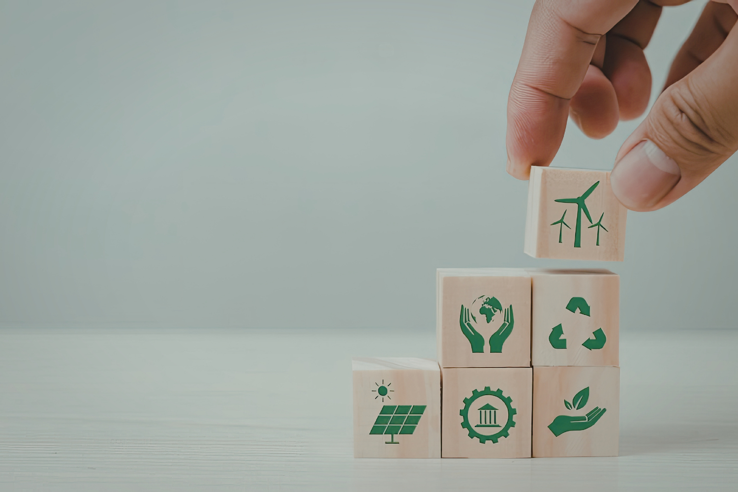 Main déposant un cube en bois illustré d’une éolienne sur une pile de cubes représentant des symboles écologiques (recyclage, énergie solaire, protection de la planète, croissance durable).